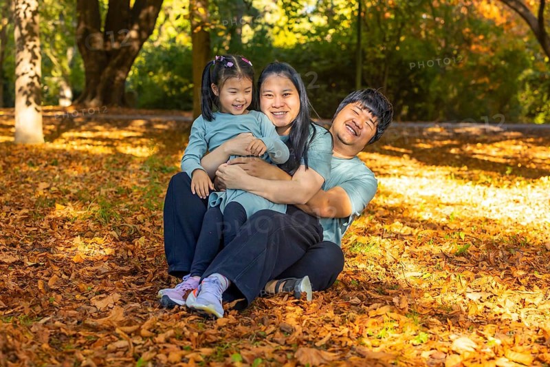 Asian family of three, smiling and embracing together, sitting on a bed of autumn leaves in a sunlit park. Warm, joyful moment capturing family bonding and outdoor enjoyment