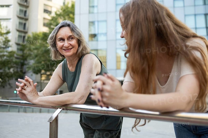 A joyful mother and daughter share quality time outdoors, leaning on a railing with a modern building backdrop. Their smiles reflect a warm and loving connection