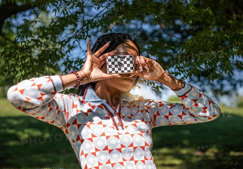A woman is joyfully capturing photos in a lush, green natural setting under a vibrant blue sky. The bright sun and warm weather enhance her outdoor photography experience