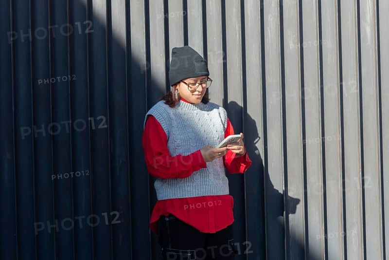 Hispanic woman wearing a beanie and glasses uses her smartphone against a modern, grey wall in an urban setting Sunlight casts a shadow, adding a dramatic effect