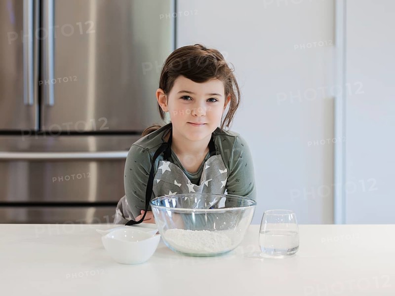 A young child, aspiring chef stands confidently at a kitchen counter with baking ingredients ready. Wearing a starry apron, the child prepares to embark on a culinary adventure