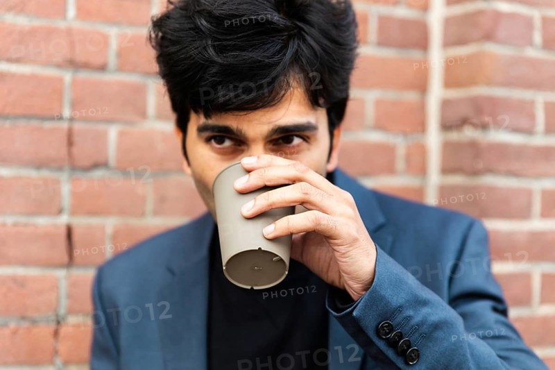 A indian businessman in a blue suit enjoys a coffee break, sipping from a cup. He stands against a brick wall during a conference of AI in Amsterdam