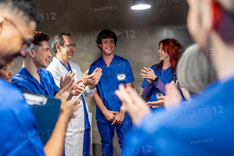 Group of diverse medical professionals, some wearing scrubs and lab coats, applauding a smiling young male intern receiving recognition and support in a hospital setting