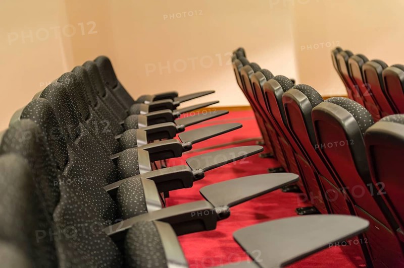 A row of empty convention center seats with black upholstery and armrests, set against a red carpet. The seating is arranged in a neatly aligned pattern, emphasizing symmetry and order