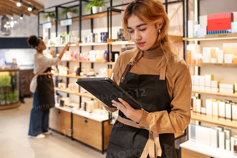 Workers in a cosmetics store are engaged in organizing products on shelves and managing inventory using a tablet. The vibrant setting emphasizes modern retail practices