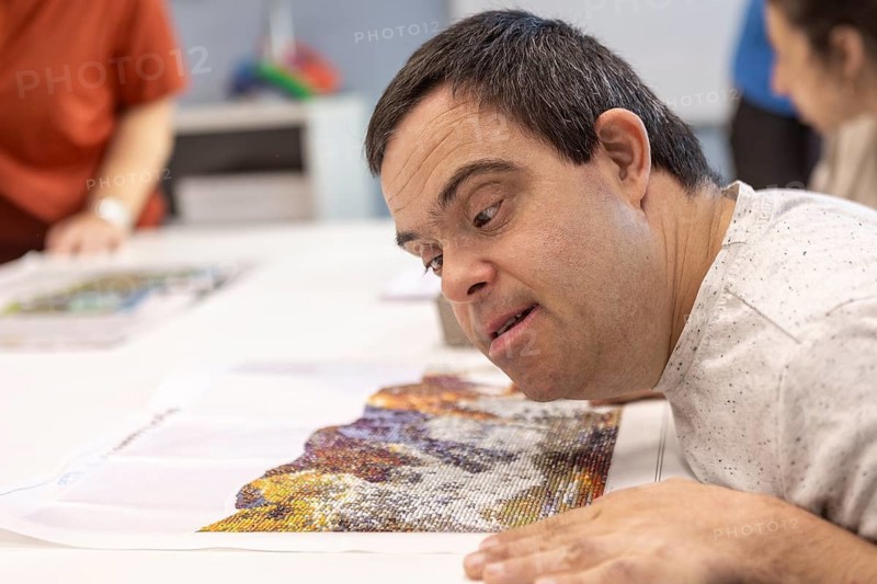 A man with Down syndrome focuses intently on a colorful mosaic artwork, carefully placing beads with precision. His expression reflects engagement, creativity, and the joy of artistic expression
