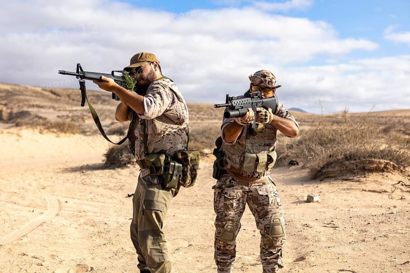 Two soldiers in tactical gear engage in a military exercise in a desert landscape, aiming their rifles forward. The dry terrain and clear sky create a stark, intense atmosphere