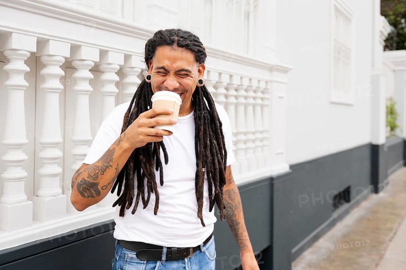 A cheerful man with dreadlocks and tattoos enjoys a coffee in Barranco, Lima, Peru He stands against a white railing, wearing casual attire, radiating positivity