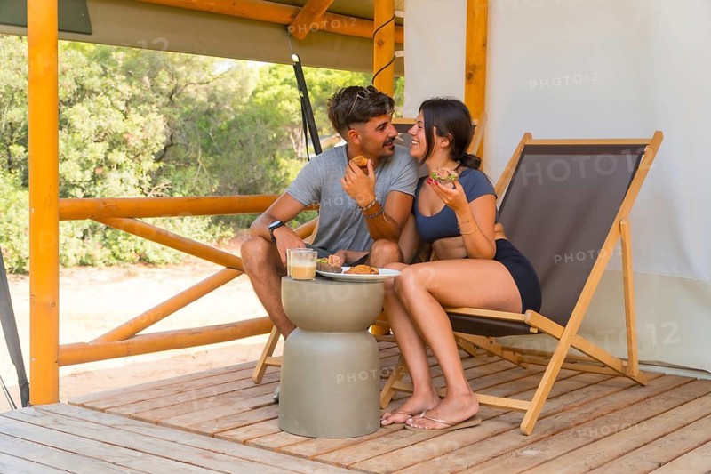 Young couple enjoying a delightful breakfast on the wooden terrace of their glamping tent, surrounded by lush nature, cherishing a romantic moment filled with love and happiness
