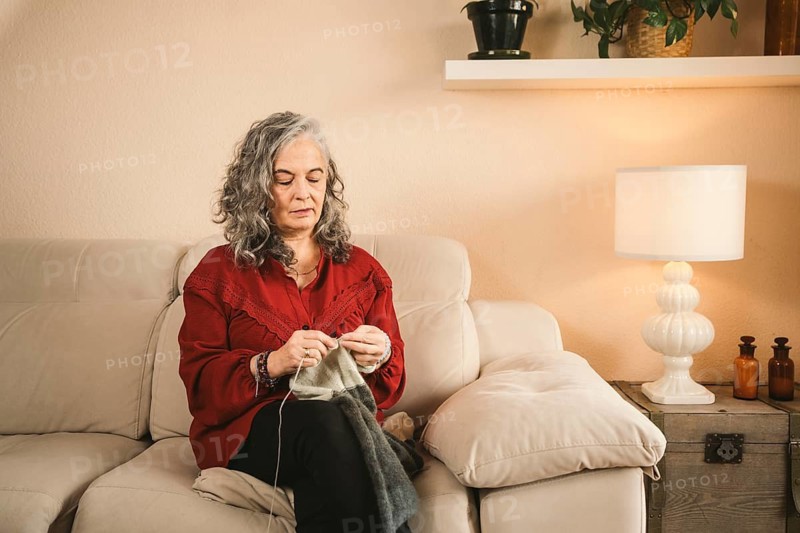 A woman with gray hair sits on a beige sofa, knitting a garment. She wears a red sweater, exuding calmness. The cozy room is softly lit by a nearby lamp