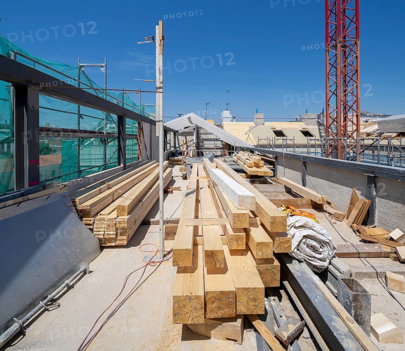 View of an active construction site featuring stacked wooden beams, scaffolding, and a crane. The clear blue sky highlights the progress and industrial environment