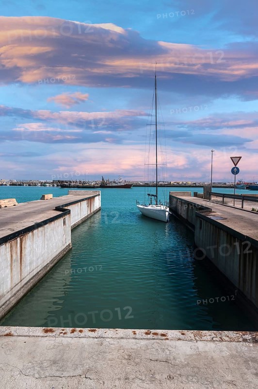 A sailboat calmly docked in a marina canal in Civitanova Marche, Italy. The sky is painted with stunning pastel colors during sunset, providing a serene backdrop