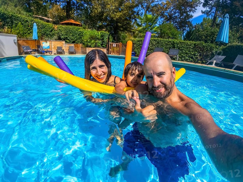 Family of three, parents and child, making a cheerful selfie together in a vibrant blue swimming pool during their summer holiday, holding colorful pool noodles and showing thumbs up for fun