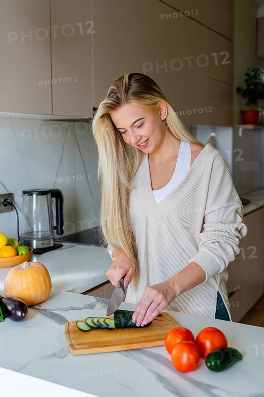 A woman in a cozy kitchen slices cucumbers on a wooden board. Surrounded by fresh vegetables, the bright space evokes a sense of home and cooking joy