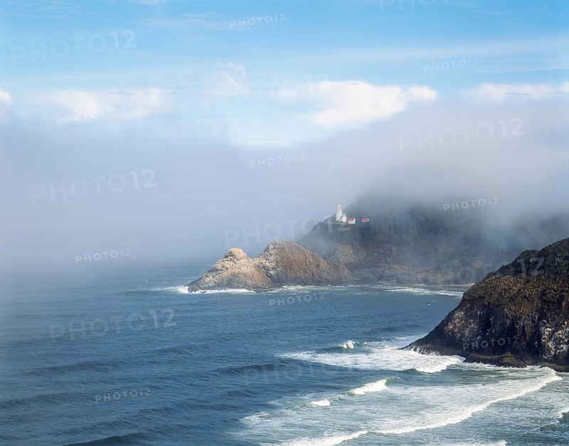 A scenic coastline featuring a lighthouse emerging from the mist atop rocky cliffs, surrounded by the ocean waves under a partly cloudy blue sky