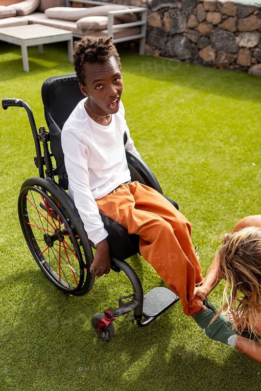 A young boy sits in a wheelchair with cerebral palsy on green grass while someone put on your shocks, emphasizing care and support in an outdoor setting. A moment of friendship