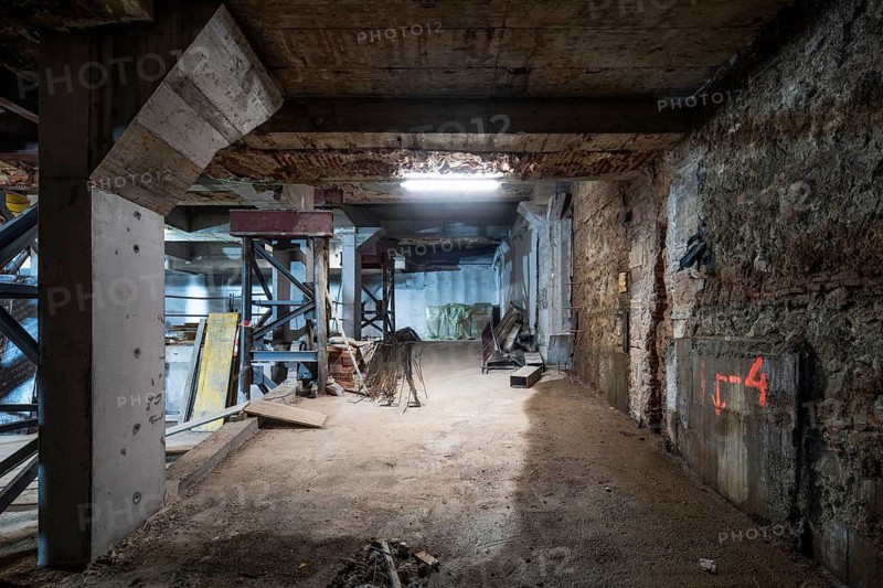 An industrial basement under construction, featuring support structures, exposed beams, scattered construction materials, and a dirt floor in progress