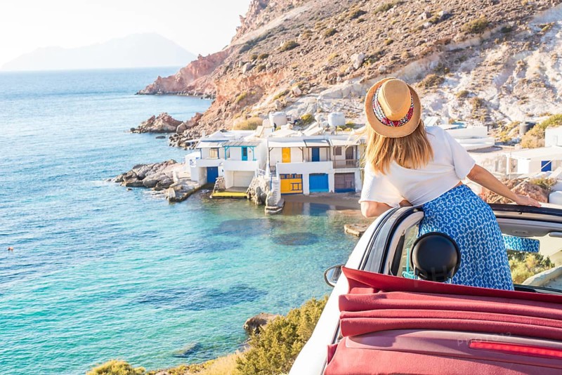 A woman in a convertible enjoys the stunning view of Fourkovouni, Milos Island. The vibrant turquoise sea and colorful coastal houses create a picturesque setting