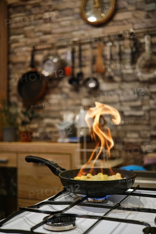 Flames are dancing from a skillet on the stove as vegetables are being sauteed in a cozy kitchen. The rustic decor features stone walls and kitchen utensils hanging in the background.