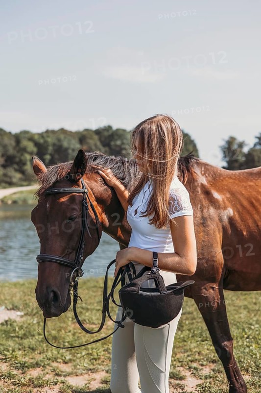 A young girl stands with her horse near a scenic lake. She gently pats the horse, creating a tranquil moment in the serene natural surroundings