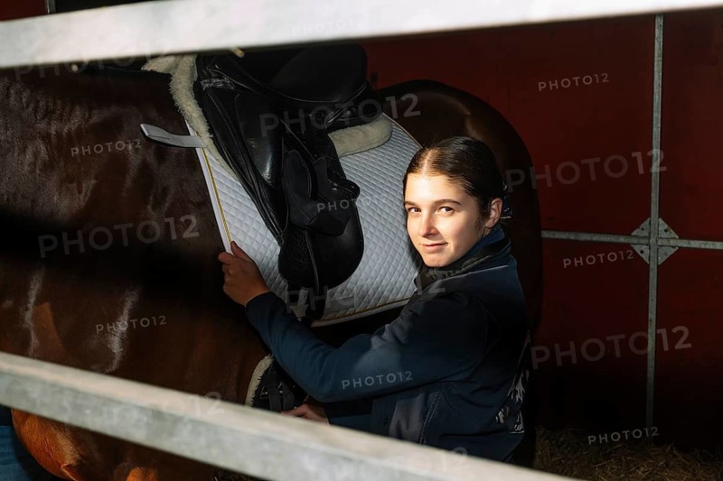 A teenage girl with a heartwarming smile adjusts a classical dressage saddle on her majestic brown horse in a stable, showcasing her care and passion for equestrian sports
