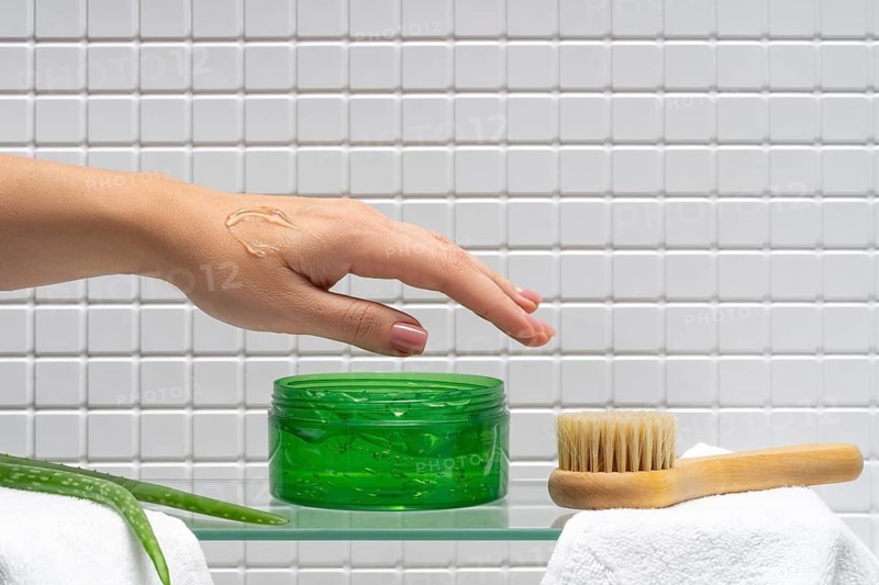 Cropped unrecognizable female hand applying body care cream, with aloe vera gel jar and wooden brush on a bathroom shelf, against a tiled wall