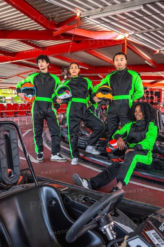 Four young adults wearing black and neon green racing suits and holding helmets, standing and sitting on karts at an indoor track, preparing for a fun and competitive go karting experience