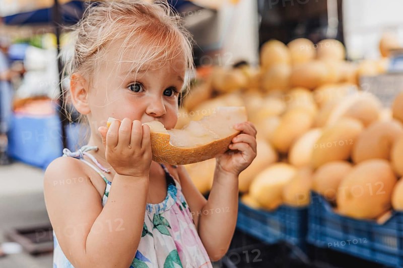 A joyful toddler savoring a fresh slice of melon at a bustling local bazaar. The vibrant market setting adds to the charm and delight of this summer scene