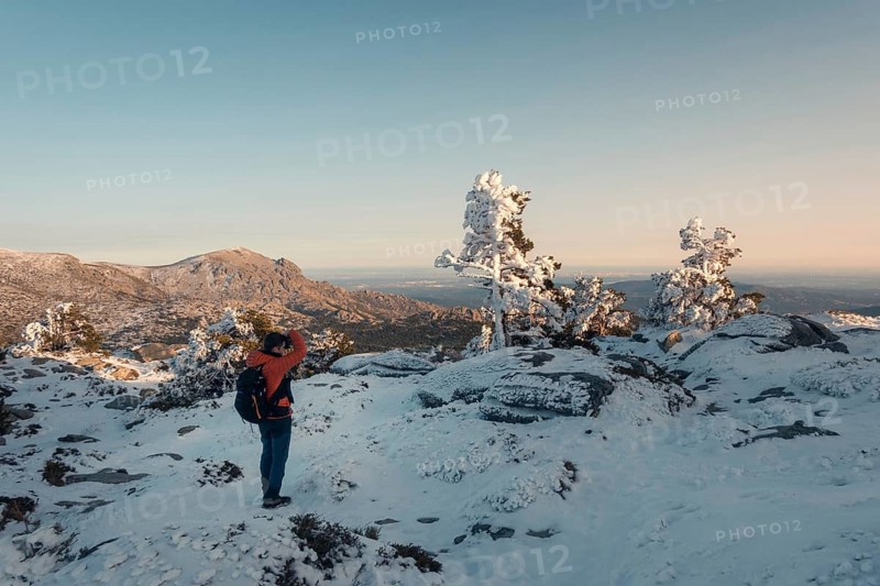 A breathtaking winter scene at Siete Picos, Guadarrama Mountains, Madrid. A hiker stands amidst snow-dusted trees and rocks, capturing the expansive mountain view