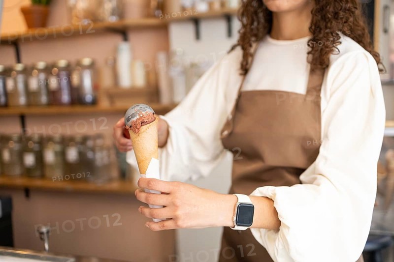 A woman clerk in a stylish brown apron serves a delicious ice cream cone in a cozy shop. The background showcases neatly organized shelves of ingredients