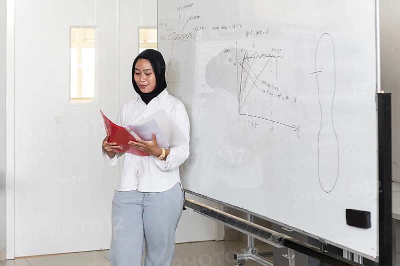 A dedicated Asian Muslim teacher with a hijab explains complex concepts to her students using a whiteboard. She holds notes and engages with the class in a modern classroom setting