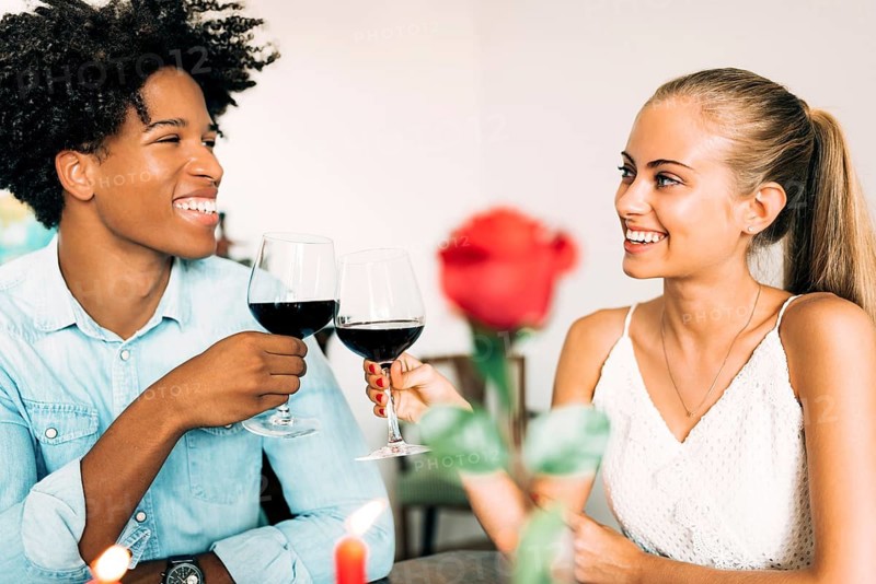 A joyful couple clinks wine glasses amid a romantic dinner setting, showcasing happiness and togetherness. A red rose adds to the intimate and cheerful ambiance