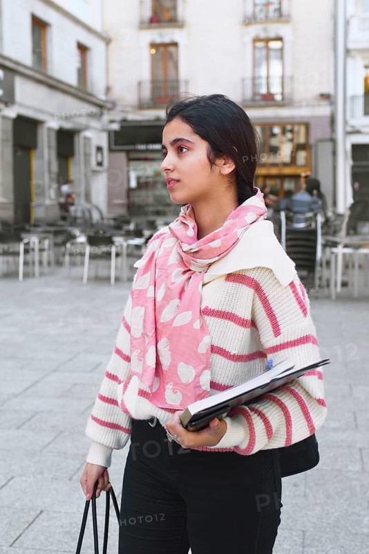 A arab student woman stands in a city street holding a notebook and tote bag. She wearing a pink and white striped sweater and a pink scarf. Background includes urban elements and architecture