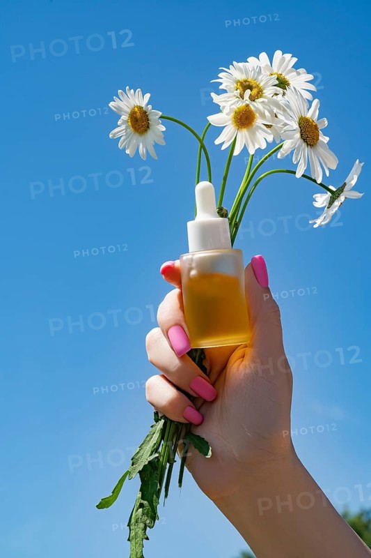 Cropped unrecognizable hand holds a cosmetic package and daisies against a vibrant blue sky. The image captures a summery, fresh vibe with a female hand slightly from below