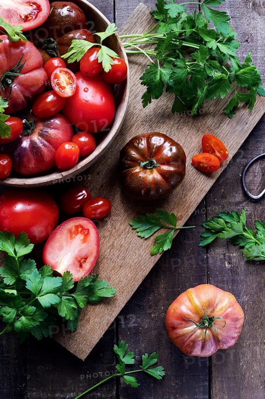 Top view of fresh tomatoes and parsley on a rustic wooden table. A wooden bowl overflows with red tomatoes, while vibrant green parsley adds a fresh touch to the scene