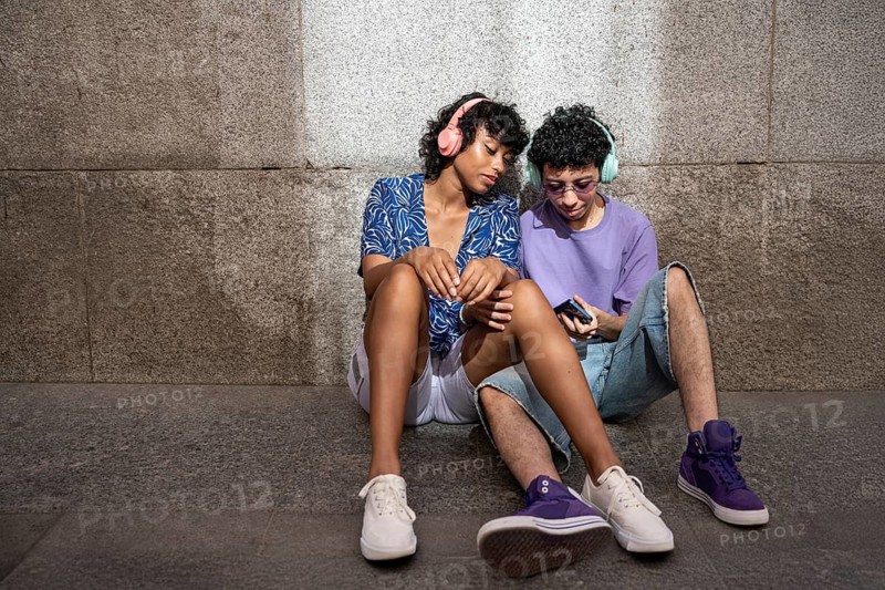 A multiethnic LGBTQ+ couple sitting against a stone wall, sharing a moment with headphones and a smartphone, dressed casually in a peaceful setting