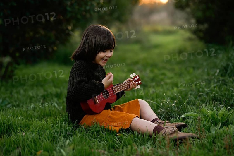 A joyful child sits on lush grass with orange trees at sunset, playing a red ukulele. The scene captures a moment of happiness, nature, and childhood innocence in the golden light