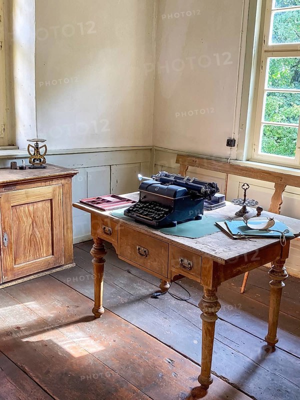 View into historical office with old historical typewriter on old small desk with turned table legs at the back left old letter scale on cupboard small cupboard in old rebuilt school house and town hall from 1830 in Open Air Museum Neuhausen, Neuhausen ob Eck, district Tuttlingen, Baden-Württemberg, Germany