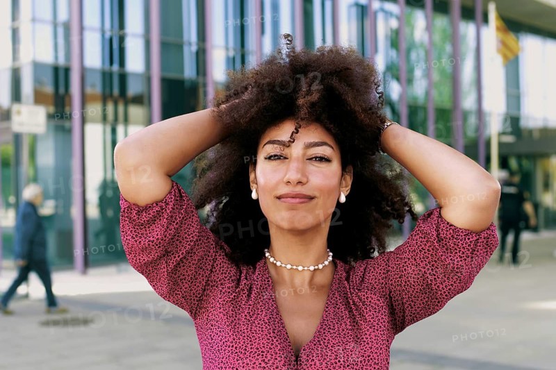 A young Arab woman with an afro hairstyle smiles confidently while posing outdoors looking at camera. She's wearing a patterned top and jewelry, standing in front of a modern glass building