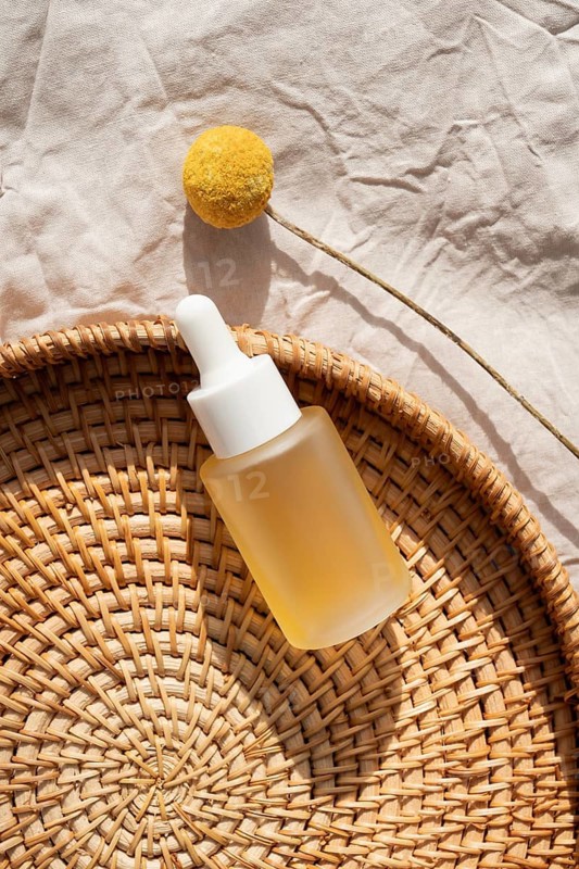 A minimalist flat lay of a cosmetic containing skincare oil, placed on a woven mat alongside a yellow billy ball flower on a textured fabric background