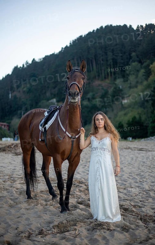 A young woman in an elegant white dress stands beside a majestic horse on a sandy beach. The serene setting is complemented by the backdrop of lush green hills