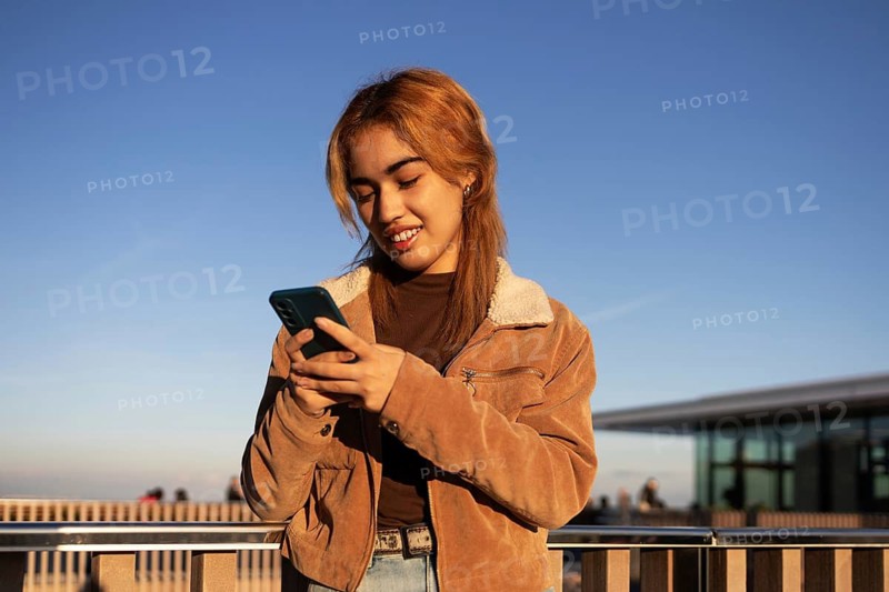 A Mixed-race woman stands outdoors using her smartphone, wearing a brown jacket and smiling The sky is clear, and she appears relaxed and content