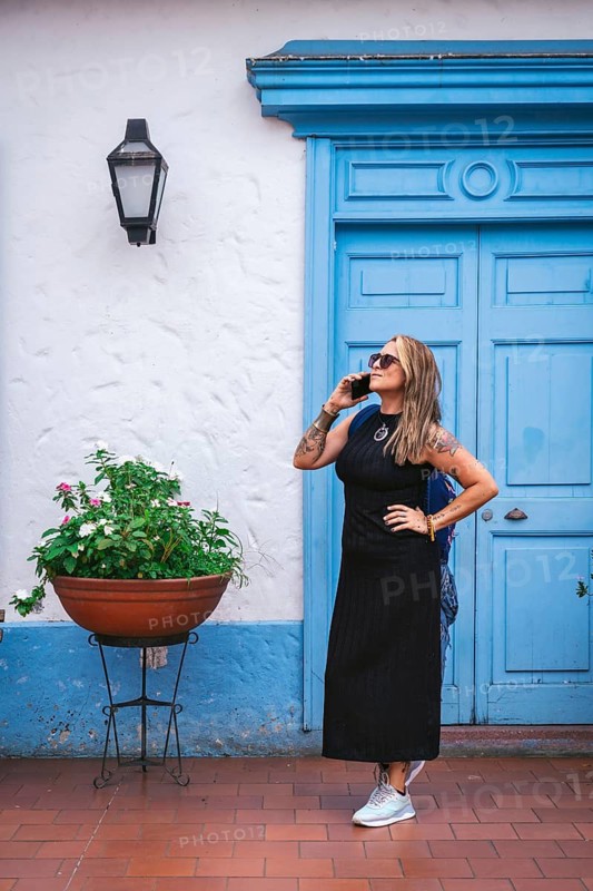 A full shot of a woman wearing a black dress and sunglasses, talking on the phone while looking up. She stands confidently in front of a blue colonial-style door, showcasing her tattoos in a vibrant urban setting.