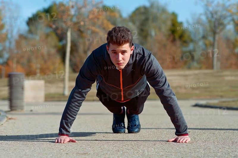 A young man performs push ups on a concrete path in a park. He is focused and dressed in sporty attire, enjoying a sunny day and outdoor exercise surrounded by nature
