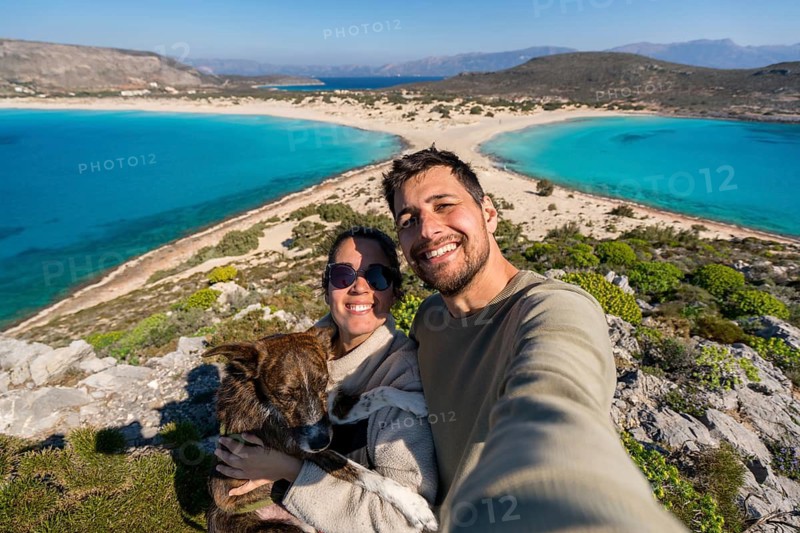 A joyful couple takes a selfie with their dog against the backdrop of a stunning, turquoise beach in Greece. The sunny day highlights the natural beauty and happy moment