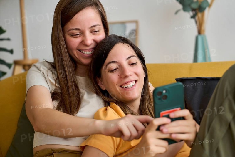 A joyful lesbian couple relaxes on a couch, sharing a happy moment while looking at a smartphone This image celebrates love and togetherness within the LGBTQIA+ community