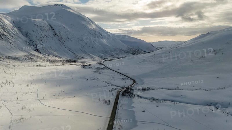 Aerial view showcasing a stunning Nordic landscape, featuring a winding road cutting through vast, snow-covered mountains under a cloudy sky. Ideal for winter travel themes, Kvaløya, Troms, Norway