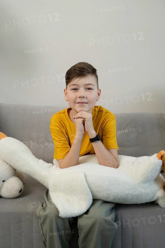 A boy wearing a yellow shirt sits on a gray sofa, resting his arms on a large white plush toy. The neutral background highlights his relaxed and thoughtful expression