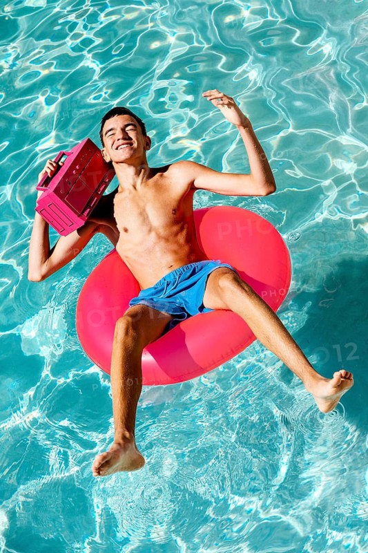 A teenage boy relaxes on a pink inflatable ring in a clear pool, holding a boombox and smiling under the bright sun. This captures a carefree summer vibe
