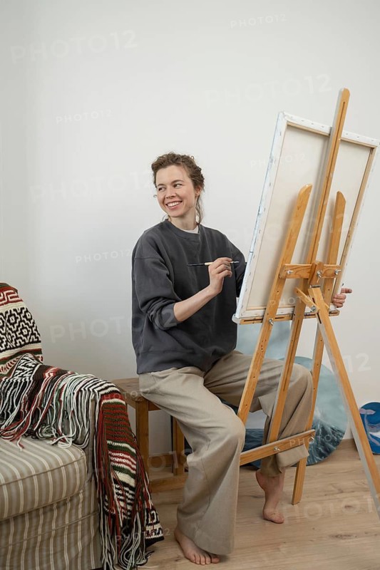Smiling artist enjoying painting at an easel in a cozy home studio. The scene captures the inspiration and joy of creativity, with a warm atmosphere and artistic decor